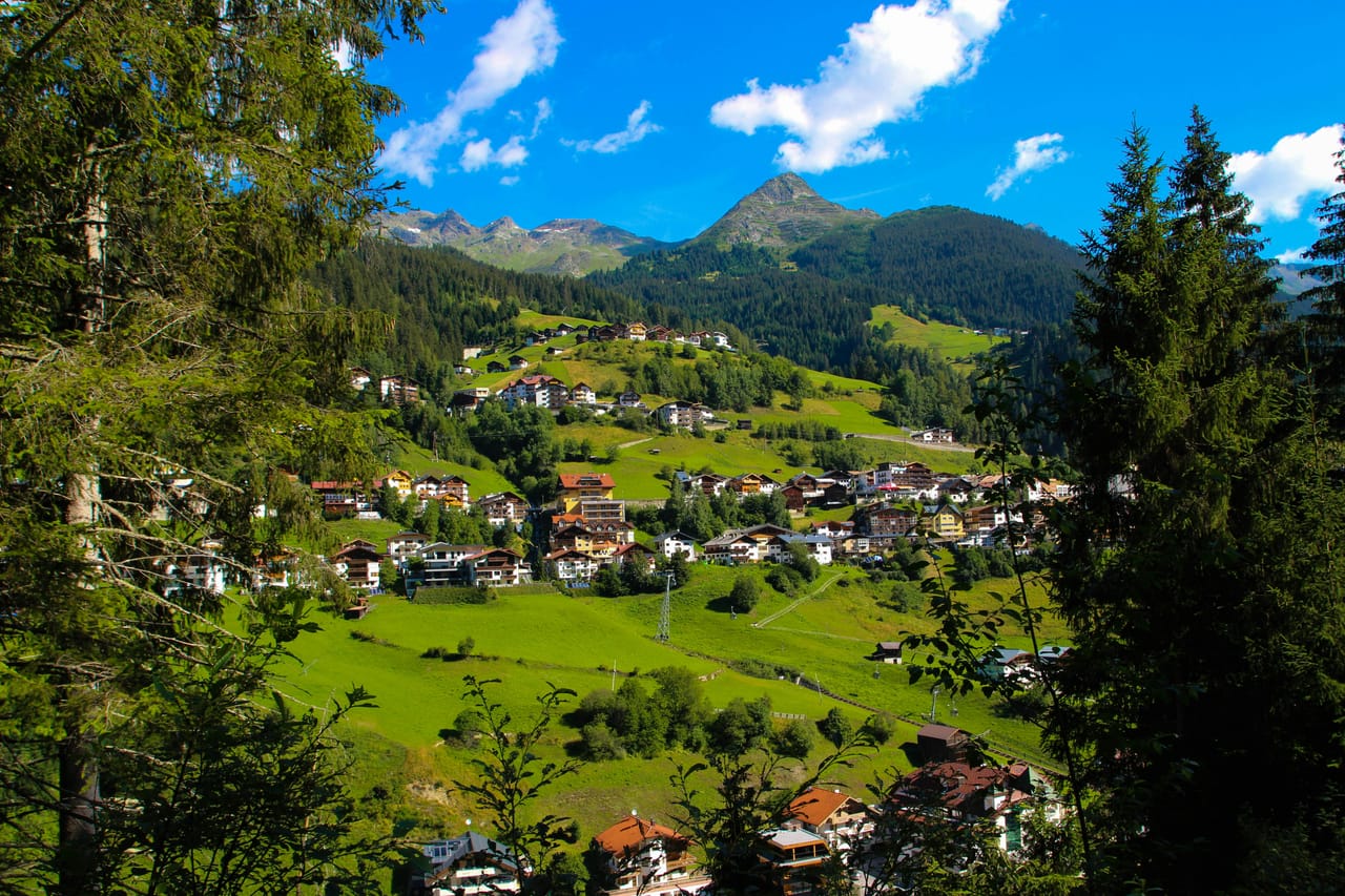 See, en el Paznaun: el pueblo sobre prados verdes, entre bosques y cumbres, bajo un cielo azul.