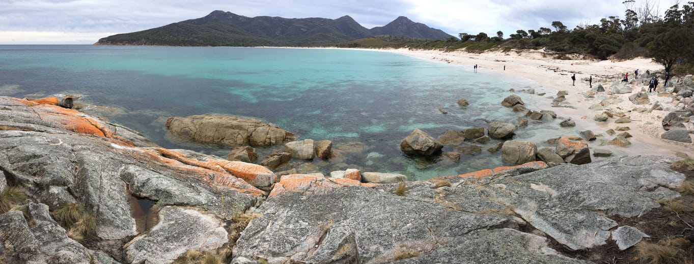 Wineglass Bay, son eau turquoise et ses rochers couverts de lichen orange, devant les montagnes de Freycinet.