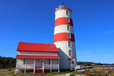 Le phare rayé de rouge de Pointe-des-Monts et la maison du gardien, sous un ciel bleu.